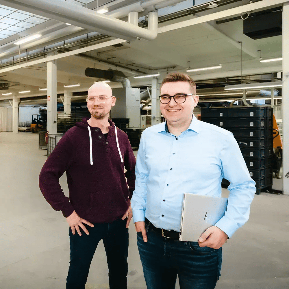 Ein IT-Mitarbeiter steht mit seinem Ansprechpartner beim Kunden in der Produktionshalle