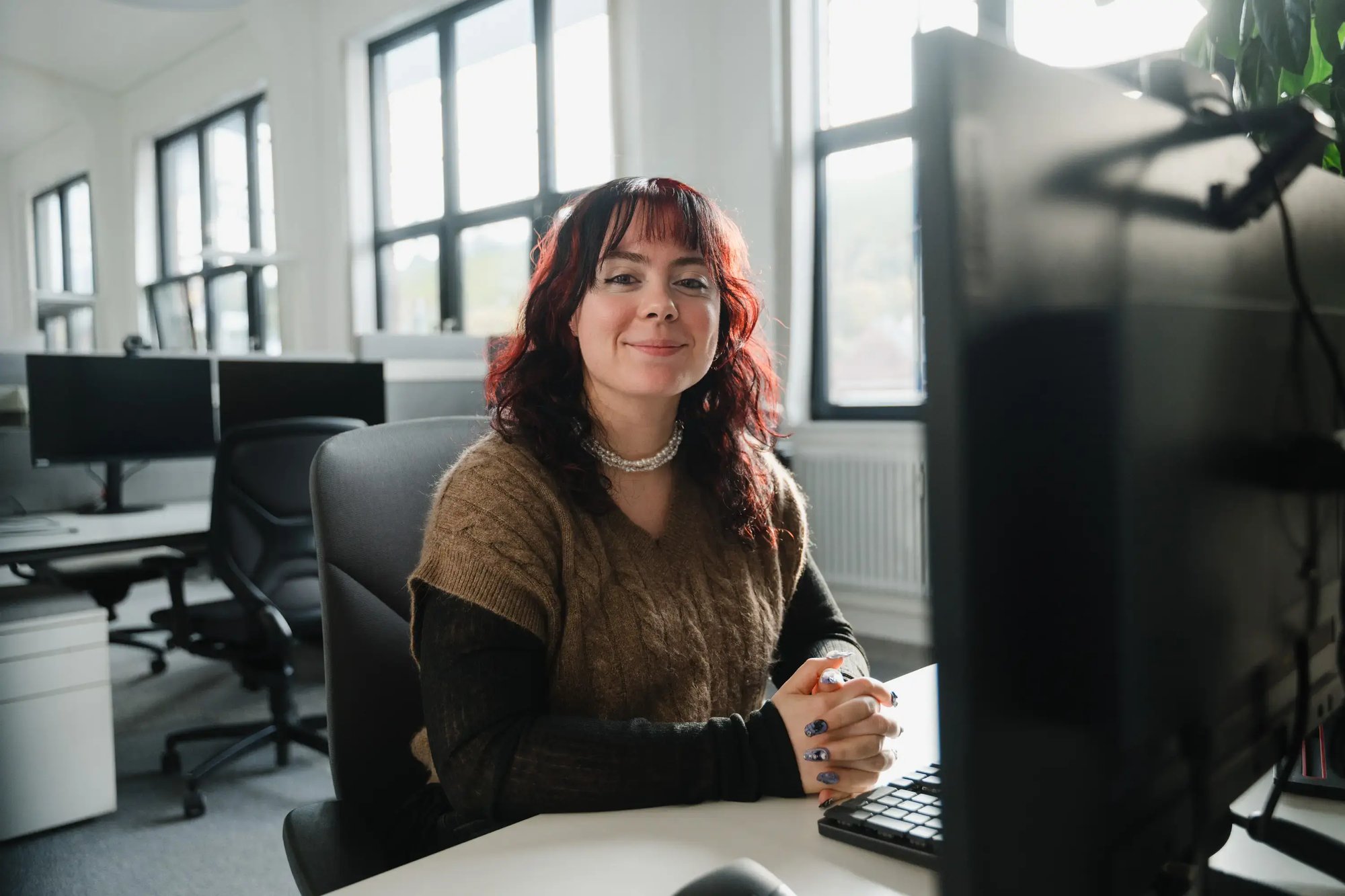 Eine junge Frau sitzt vor einem Monitor und lächelt in die Kamera